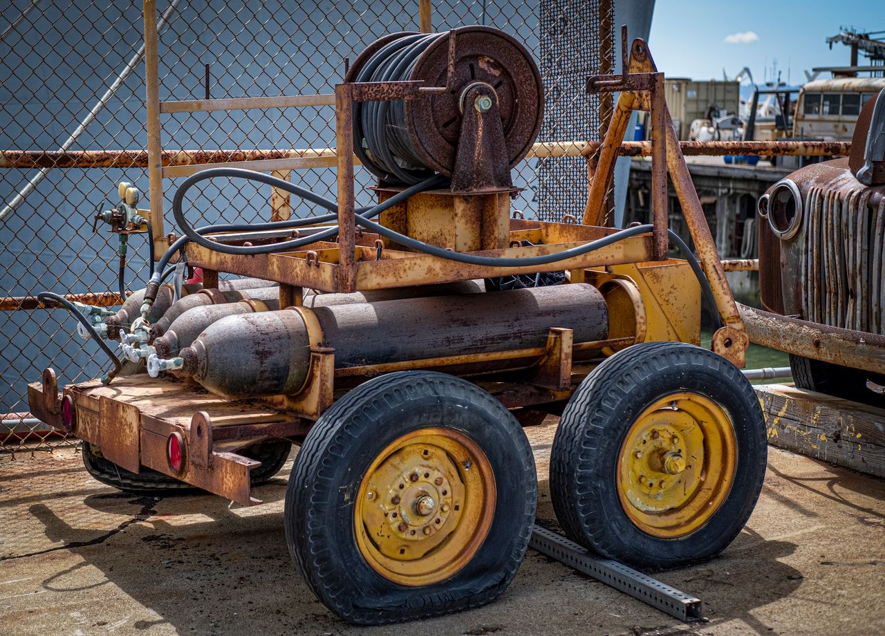 why-choose-us-01 Old rusty industrial cart with hoses and tanks, parked on a sunlit day.